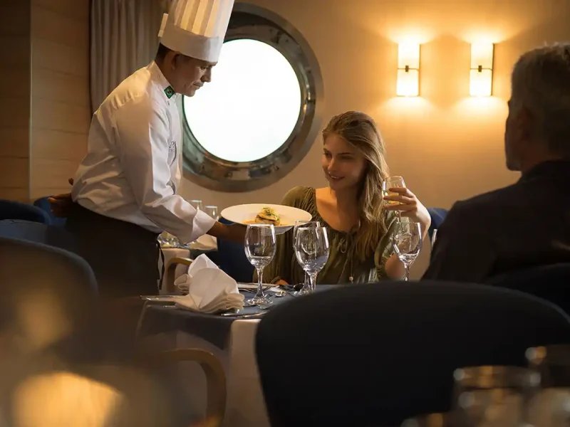 A chef serving a plated dish to guests in the dining room aboard La Pinta